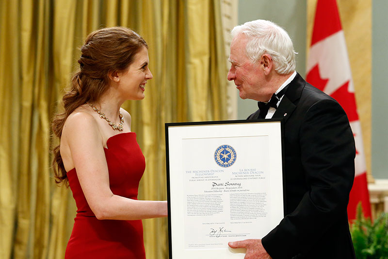 A smiling woman in a red dress receives a certificate from a white haired man in a black tuxedo.