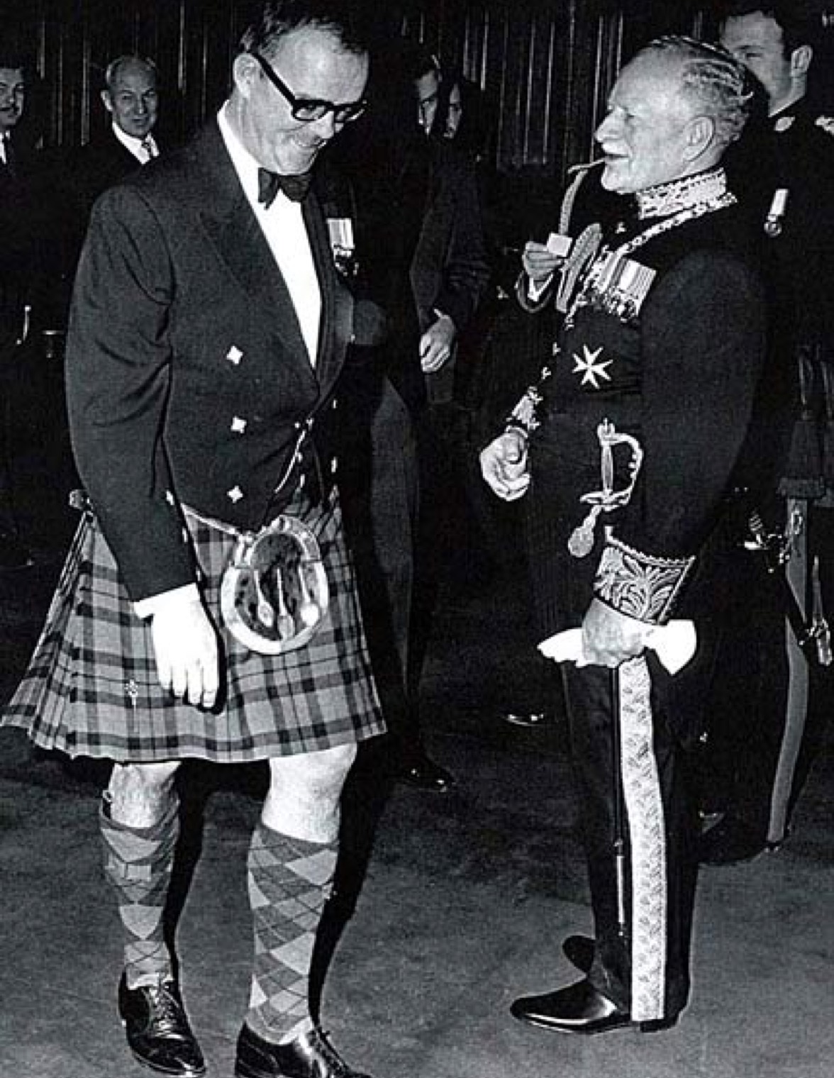 Two men are in the foreground. The man on the left is wearing a kilt, the man on the right is wearing a military uniform.