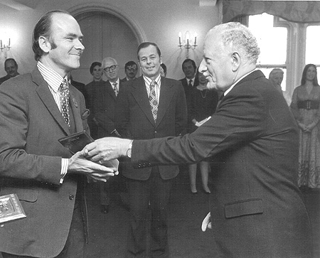 A black and white image of a white-haired man offering an award to another man. Both are wearing suits.