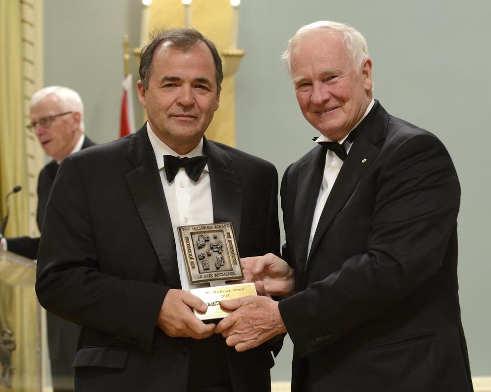 A man presents a large award to another man, both in tuxedos.